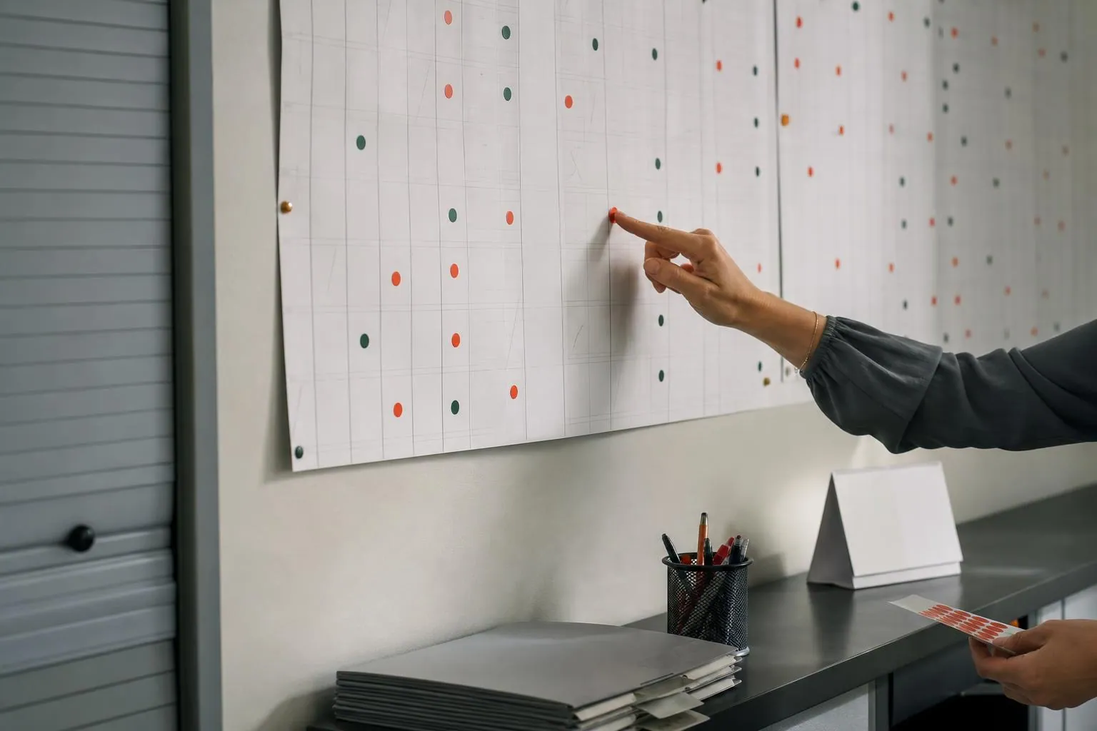 Un planning annuel en bandes de papier punaisé au mur d’un bureau administratif, une main y posant une pastille adhésive de couleur